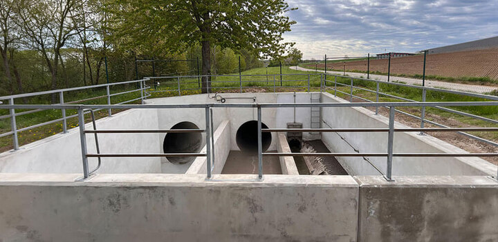 Offenes Betonbauwerk mit zwei großen Rohrzuläufen, umlaufendem Geländer und einer Leiter, umgeben von Bäumen und Feldern.